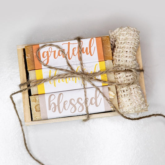 A wooden crate with 3 bar soaps and a fabric soap saver pouch, tied together with twine. Crate contains three bar soaps labeled 'grateful', 'thankful', and 'blessed'.
