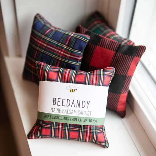 A group of 4 fabric pillows with various plaid patterns, on white windowsill. The pillow in front has a white and green label.