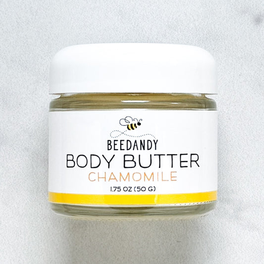 Beedandy Chamomile Body Butter in a small clear glass jar with a white lid, shown on a light gray background. The label features a bee logo and reads “Body Butter, Chamomile, 1.75 oz (50 g),” with a yellow accent band at the bottom.