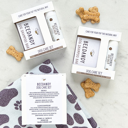 The image shows two packages of the Dog Care Set, each containing a dog balm and a full size shampoo bar, packaged in white boxes with branding and logos. There are also treats visible beside the boxes on a patterned fabric.