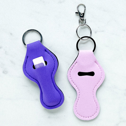 Two fabric lip balm holders with key rings, one dark purple holding a lip balm tube and one light purple shown empty, on a light background.