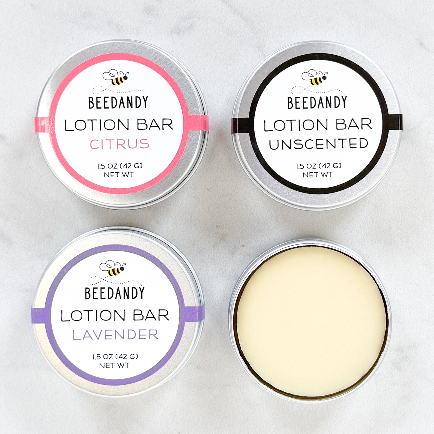 Four round metal tins of Beedandy lotion bars, including citrus, unscented, and lavender varieties, shown from above, with one tin open to display the lotion bar.