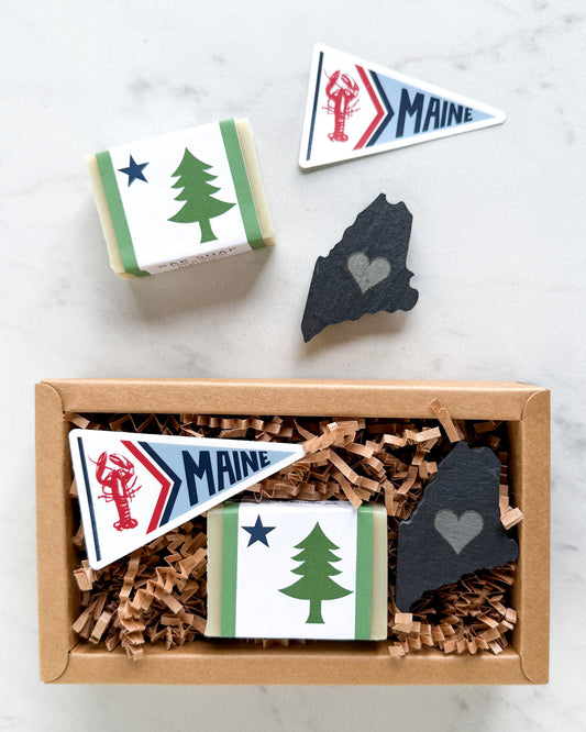 A gift box containing Maine-themed items, including a Maine pennant, a wrapped bar with a pine tree design, and a slate Maine-shaped ornament, shown from above with one box open and one closed. Another set of items beside box on white background.