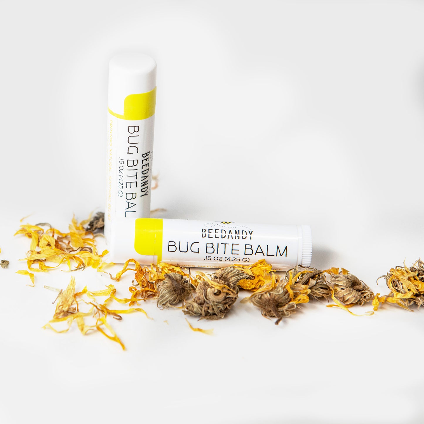 Two small white tubes with yellow labeling labeled Bug Bite Balm. One is standing up, one is lying flat. All on a white background with a small pile of calendula flowers.