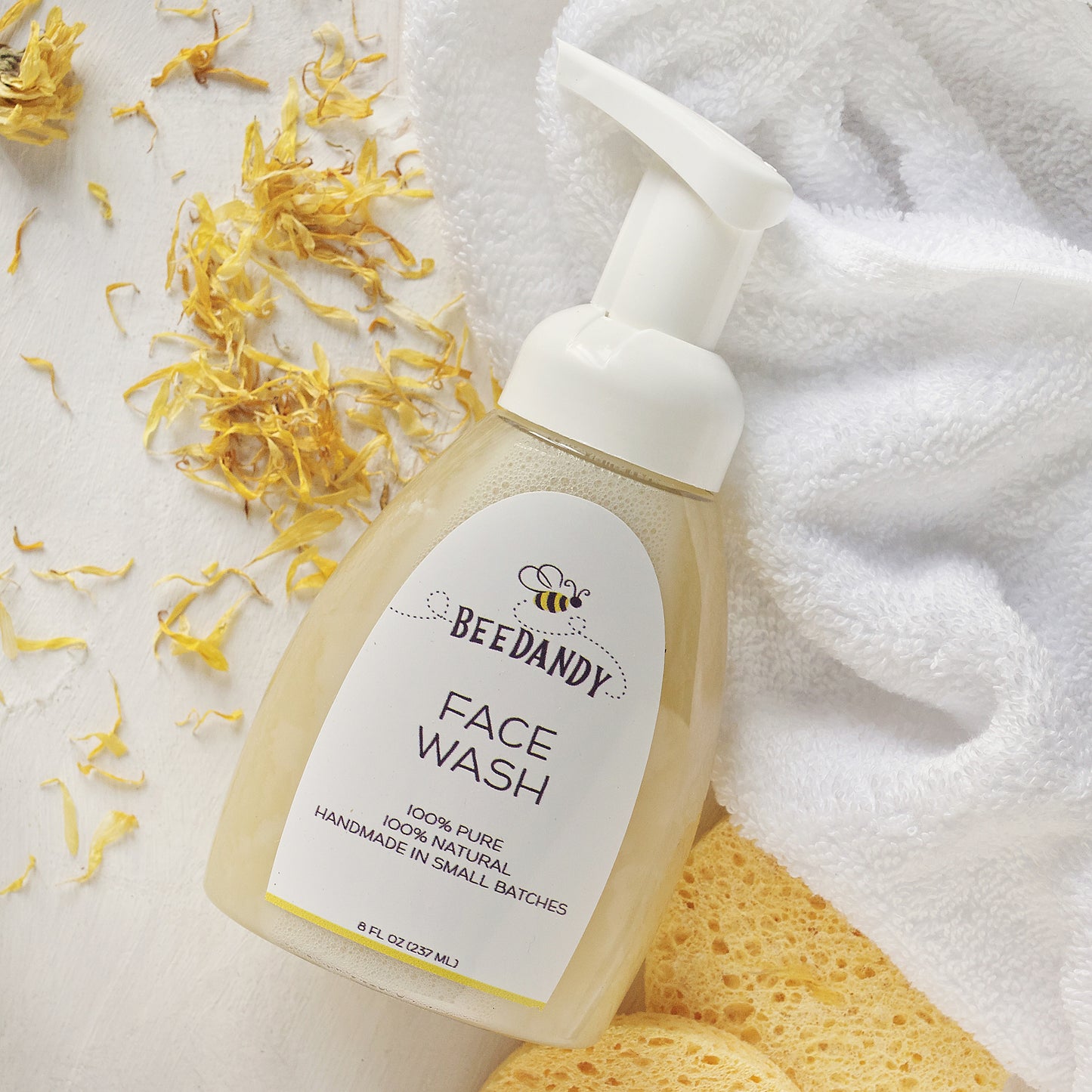 A pump bottle of face wash with a white label, placed on a surface with a white towel and scattered flower petals around it.