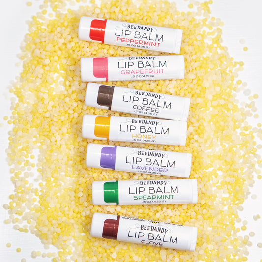 Seven Beedandy lip balm tubes in different flavors displayed on a bed of yellow beeswax pellets.