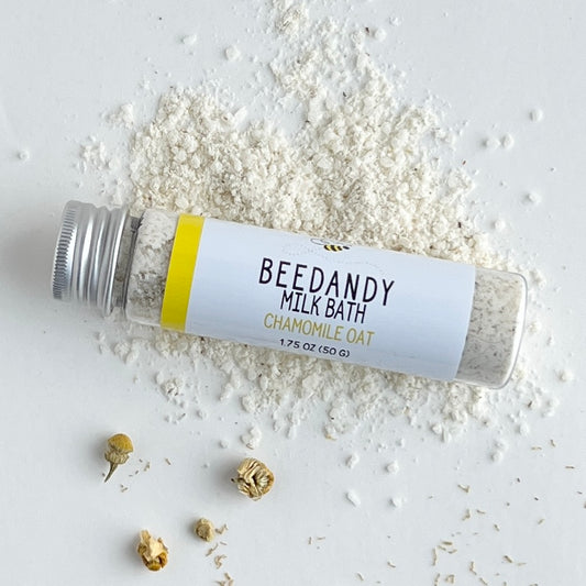 A clear tube of milk bath labeled chamomile oat with white label and silver cap. Against a light grey background and light yellow powder.