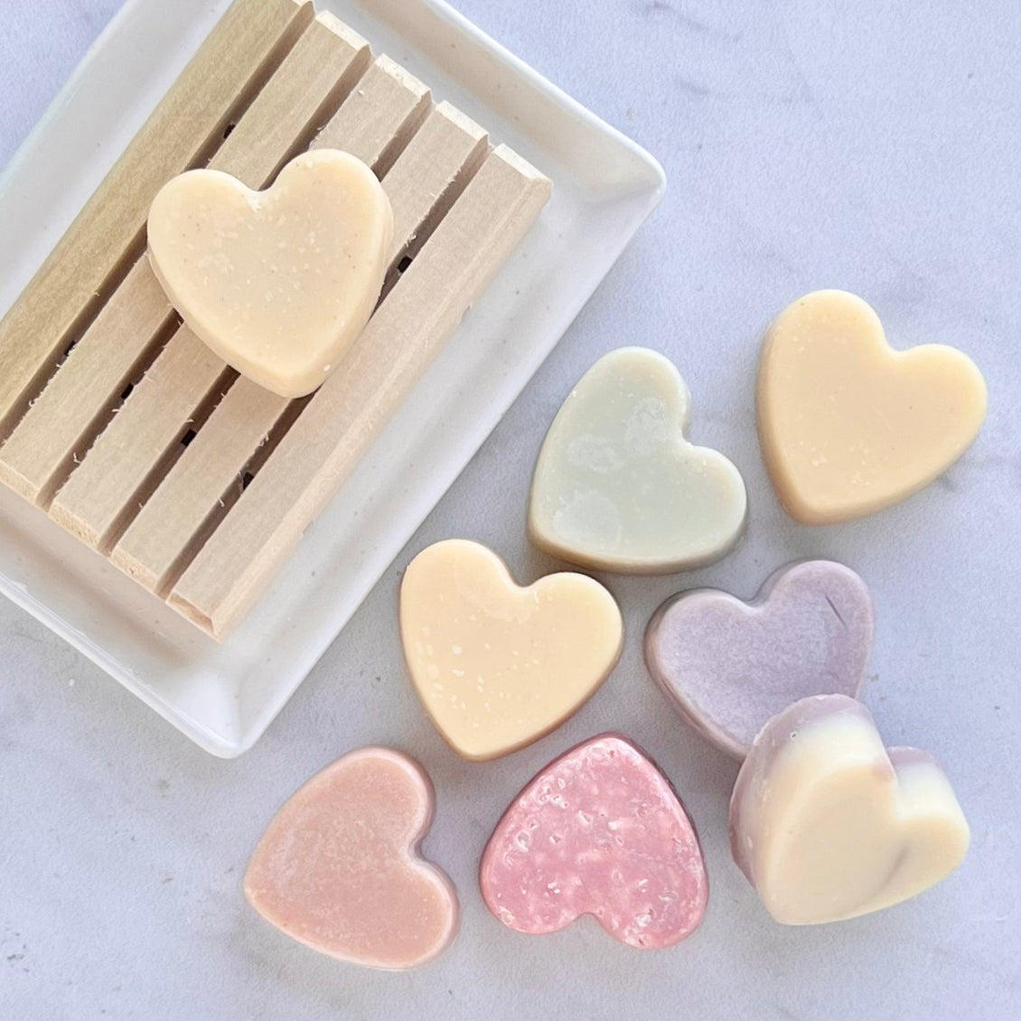 Assorted mini heart-shaped soaps in soft pastel colors (cream, pink, lavender, and mint) arranged on a white surface, with one soap resting on a wooden soap dish.