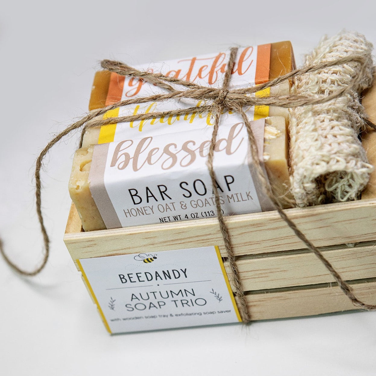 A wooden crate with 3 bar soaps and a fabric soap saver pouch, tied together with twine. Crate contains three bar soaps labeled 'grateful', 'thankful', and 'blessed'. Crate has a white and yellow label stating "Autumn Soap Trio".