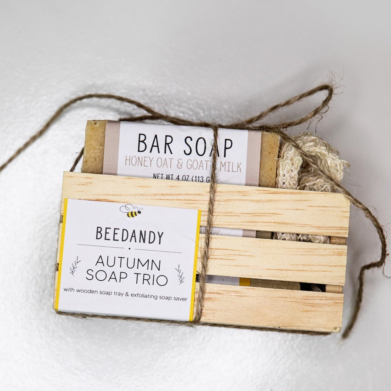 A wooden crate with 3 bar soaps and a fabric soap saver pouch, tied together with twine. Crate contains three bar soaps labeled 'grateful', 'thankful', and 'blessed'. Crate has a white and yellow label stating "Autumn Soap Trio with wooden soap tray and exfoliating soap saver"