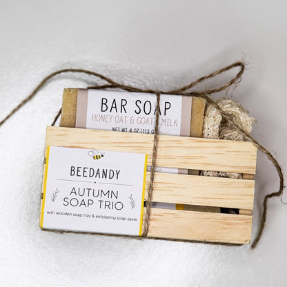 A wooden crate with 3 bar soaps and a fabric soap saver pouch, tied together with twine. Crate contains three bar soaps labeled 'grateful', 'thankful', and 'blessed'. Crate has a white and yellow label stating "Autumn Soap Trio with wooden soap tray and exfoliating soap saver"