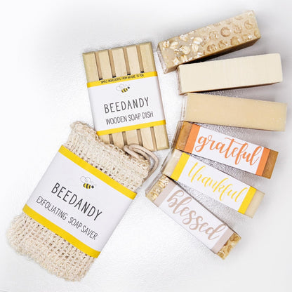 Fabric exfoliating soap saver, wooden soap dish, three unlabeled soaps and three bar soaps labeled 'grateful', 'thankful', and 'blessed'. All against a white background.
