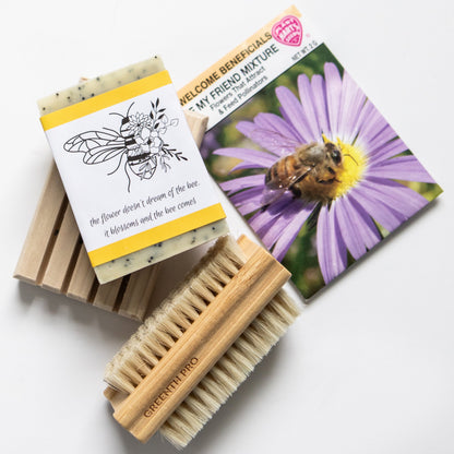 Eco-friendly flat lay featuring a natural handmade soap bar with botanical bee design and quote “the flower doesn’t dream of the bee. it blossoms and the bee comes,” a packet of pollinator-friendly flower seeds showing a bee on a purple daisy, a 2 sided bamboo scrub brush and a wooden soap dish on a white background. Sustainable self-care and zero-waste lifestyle concept with natural bath accessories and bee conservation theme.