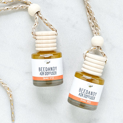 2 glass air diffusers with wooden lids, attached to braided ropes, labeled with white and orange label "Orange Spice"