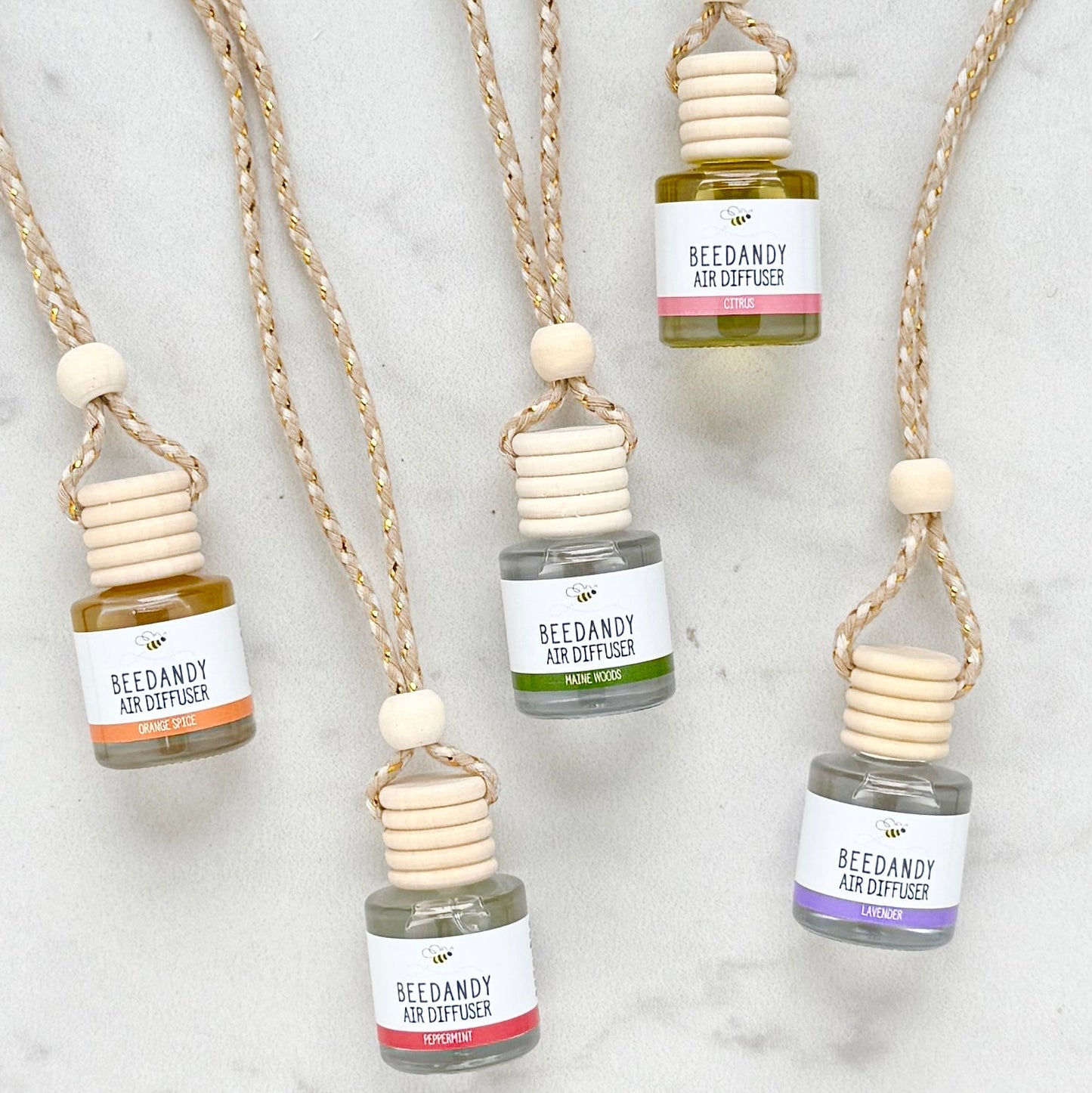 A collection of 5 glass air diffusers with wooden lids, attached to braided ropes. 5 scents with various colored labels. Scents left to right are orange spice, peppermint, Maine woods, citrus, lavender.  