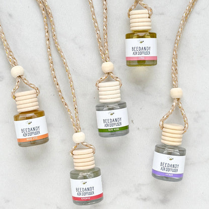 A collection of 5 glass air diffusers with wooden lids, attached to braided ropes. 5 scents with various colored labels. Scents left to right are orange spice, peppermint, Maine woods, citrus, lavender.  