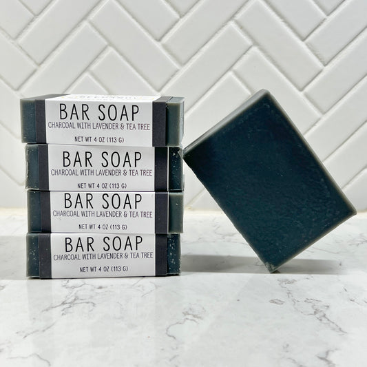 A stack of 4 bar soaps with black and white label labeled "Charcoal with Lavender & Tea Tree" with a fifth unlabeled bar on the right leaning against the stack. Soaps are black in color. On white marble counter with white tile backsplash.