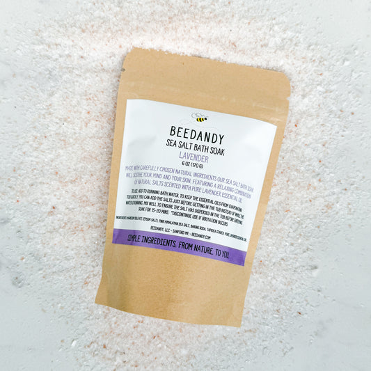 A brown Kraft pouch of BEEDANDY sea salt bath soak with a label listing natural ingredients such as Epsom salt and lavender essential oil. Pouch is against a background of pink salt. 