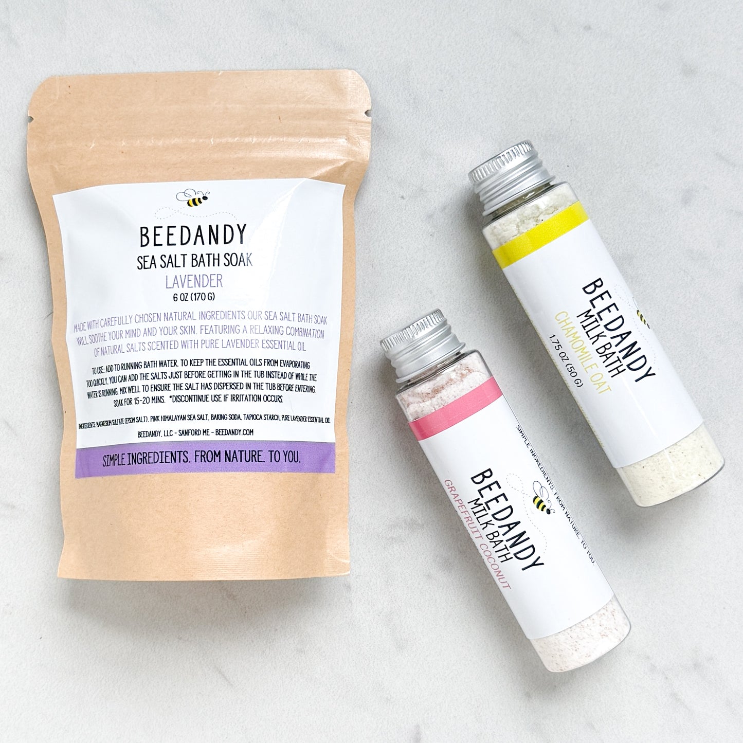 A Beedandy lavender sea salt bath soak in a kraft pouch alongside two Beedandy milk bath bottles on a light background.
