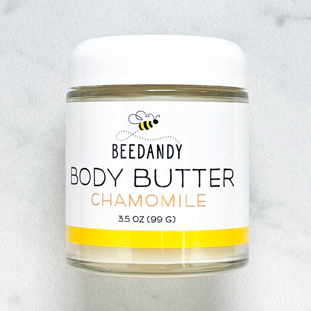Beedandy Chamomile Body Butter in a small clear glass jar with a white lid, shown on a light gray background. The label features a bee logo and reads “Body Butter, Chamomile, 3.5 oz (99 g),” with a yellow accent band at the bottom.