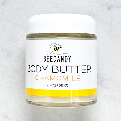 Beedandy Chamomile Body Butter in a small clear glass jar with a white lid, shown on a light gray background. The label features a bee logo and reads “Body Butter, Chamomile, 3.5 oz (99 g),” with a yellow accent band at the bottom.