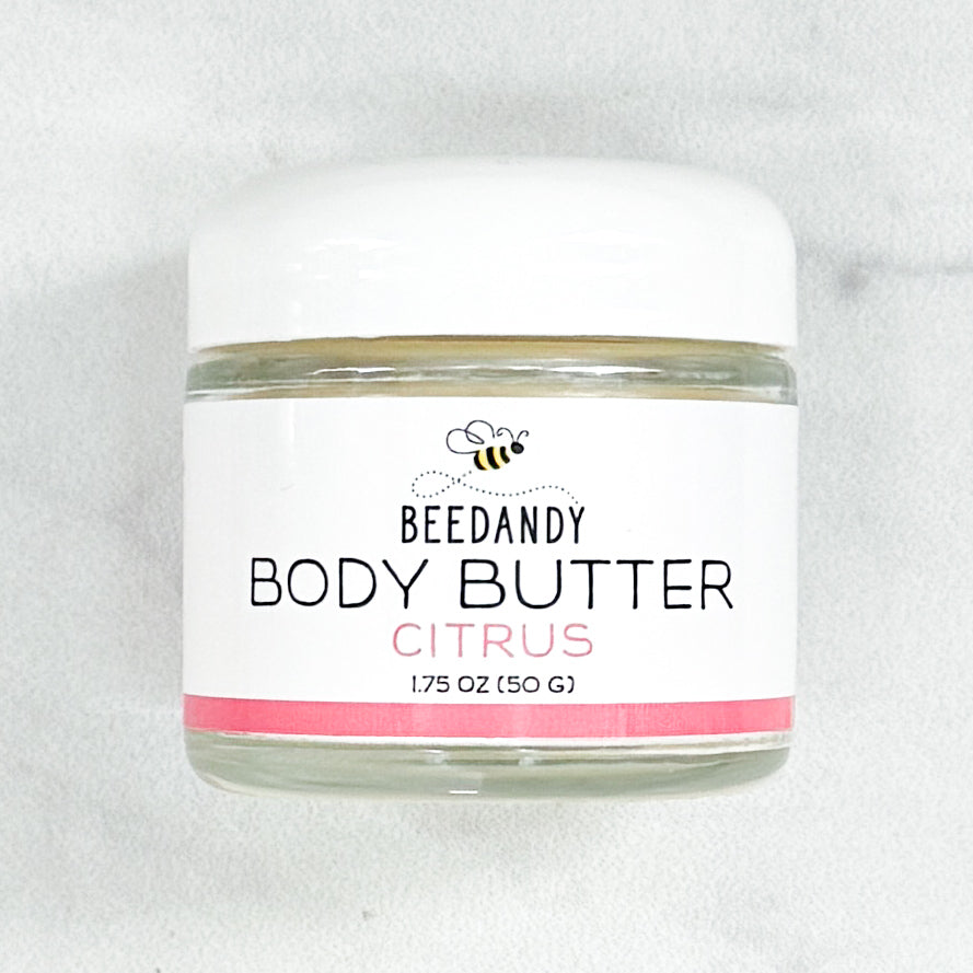 Beedandy Citrus Body Butter in a small clear glass jar with a white lid, shown on a light gray background. The label features a bee logo and reads “Body Butter, Citrus, 1.75 oz (50 g),” with a pink accent band at the bottom.