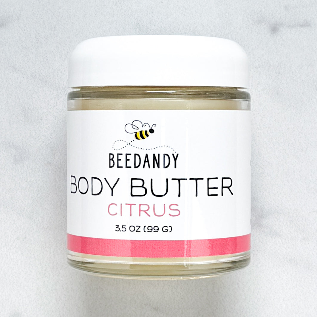 Beedandy Citrus Body Butter in a small clear glass jar with a white lid, shown on a light gray background. The label features a bee logo and reads “Body Butter, Citrus, 3.5 oz (99 g),” with a pink accent band at the bottom.