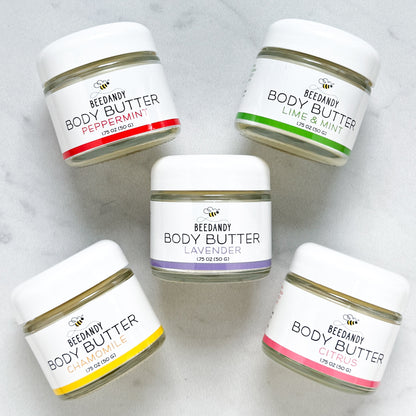 Five small glass jars of Beedandy Body Butter arranged in a circular pattern on a light gray surface. Each jar has a white lid and label with a bee logo. Scents shown are Peppermint (red), Lime & Mint (green), Lavender (purple), Chamomile (yellow), and Citrus (pink), each labeled 1.75 oz (50 g).