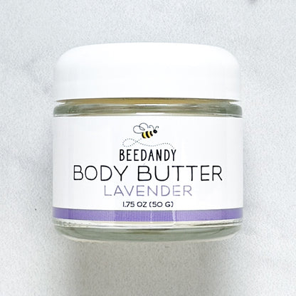 Beedandy Lavender Body Butter in a small clear glass jar with a white lid, shown on a light gray background. The label features a bee logo and reads “Body Butter, Lavender, 1.75 oz (50 g),” with a purple accent band at the bottom.