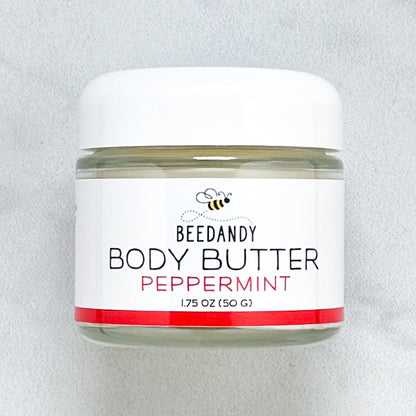 Beedandy Peppermint Body Butter in a small clear glass jar with a white lid, shown on a light gray background. The label features a bee logo and reads “Body Butter, Peppermint, 1.75 oz (50 g),” with a red accent band at the bottom.