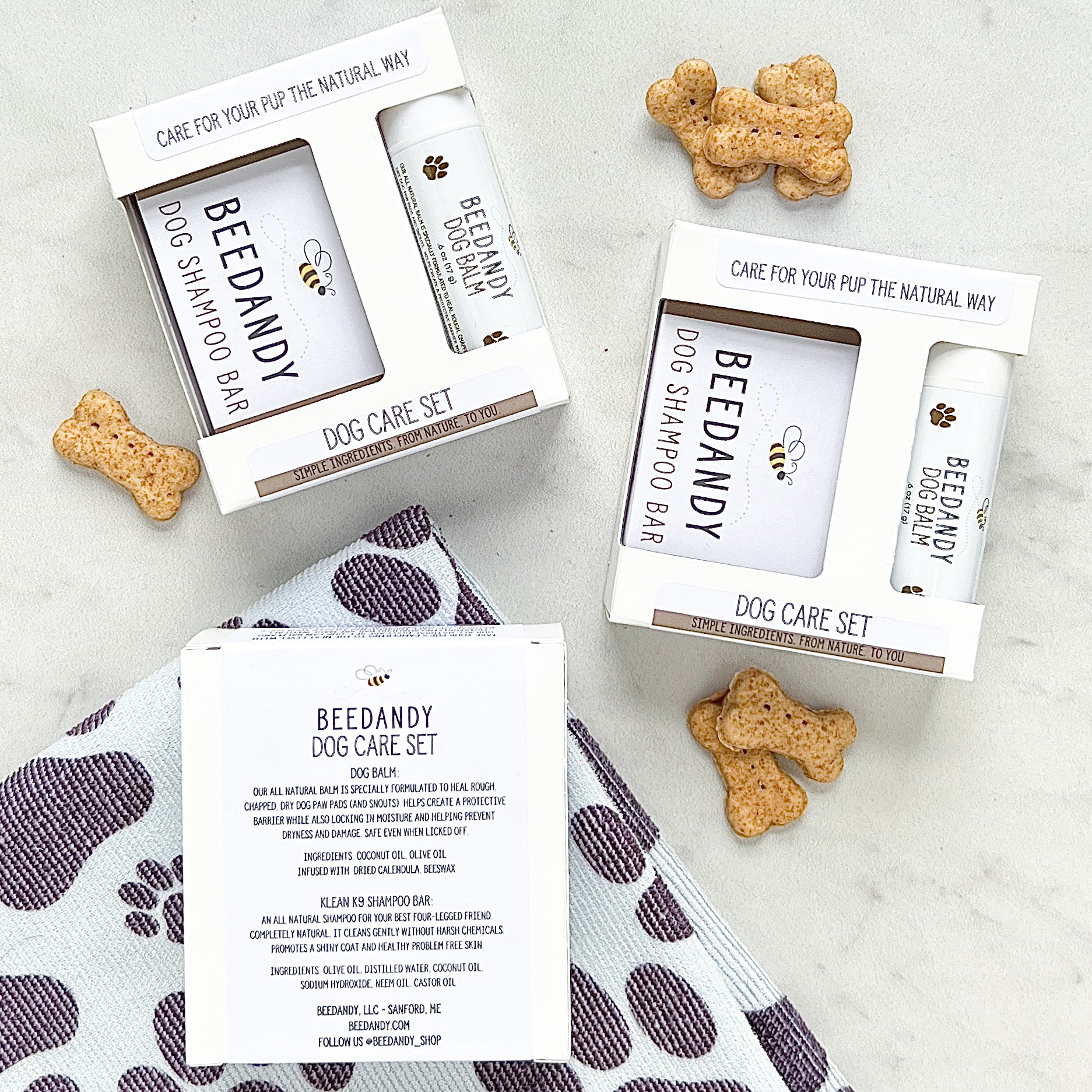 The image shows two packages of the Dog Care Set, each containing a dog balm and a full size shampoo bar, packaged in white boxes with branding and logos. There are also treats visible beside the boxes on a patterned fabric.