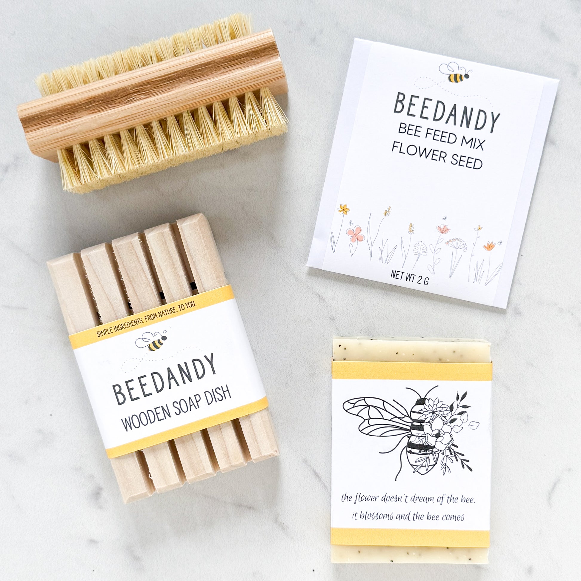 Items arranged in a square include a wooden nail brush, packet of flower seed, bar soap with bee label and wooden soap dish, all against a white marble background.
