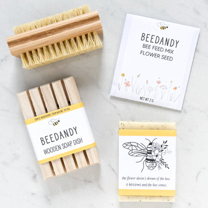 Items arranged in a square include a wooden nail brush, packet of flower seed, bar soap with bee label and wooden soap dish, all against a white marble background.
