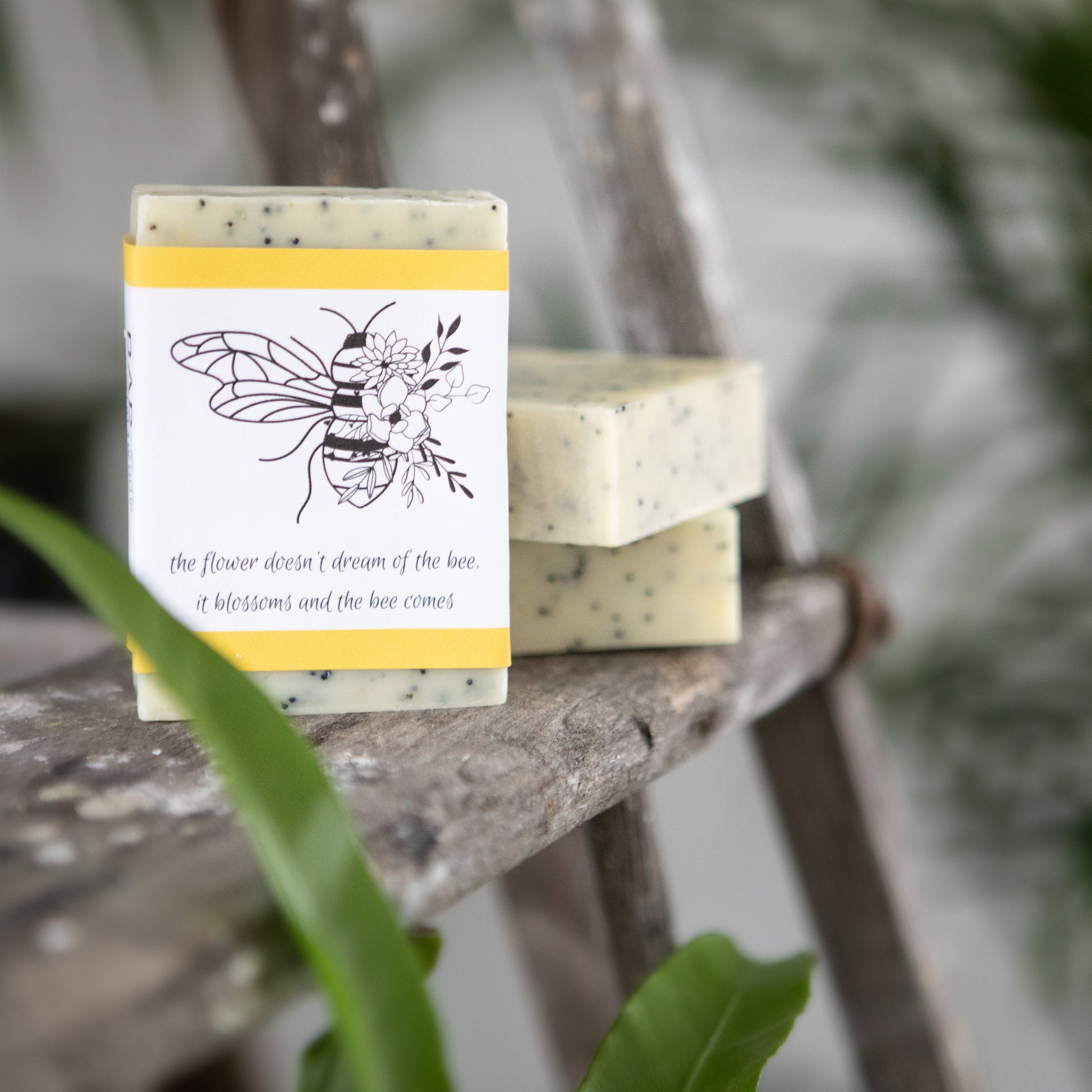 Handmade soap bar with poppy seeds, wrapped with a bee illustration label, displayed outdoors on a wooden surface.