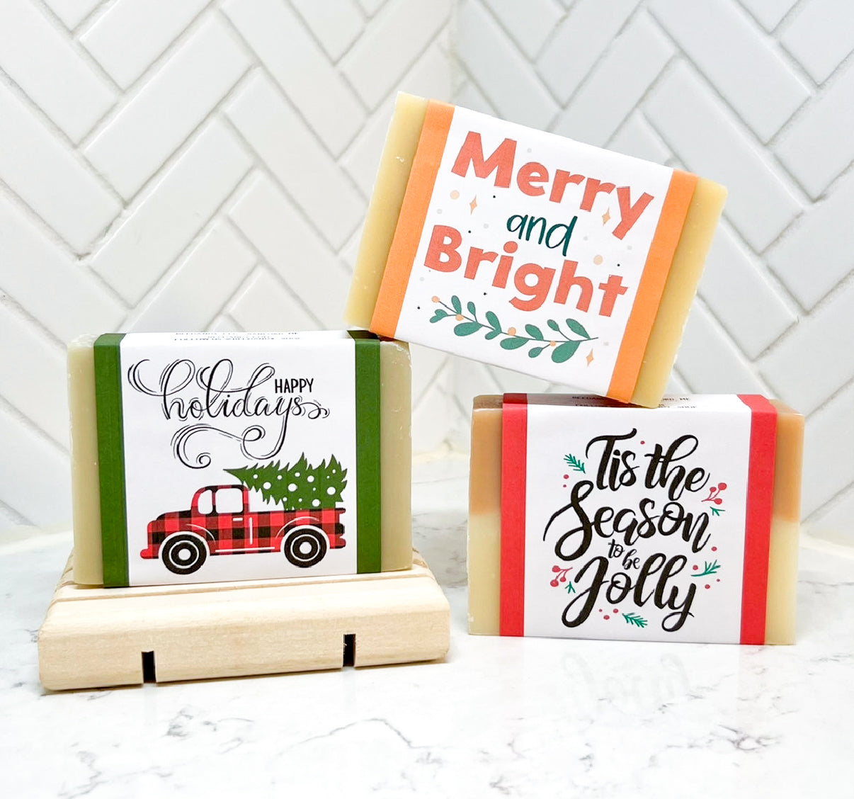 Three holiday-themed bar soaps with decorative labels displayed on a light surface, one resting on a wooden soap dish.