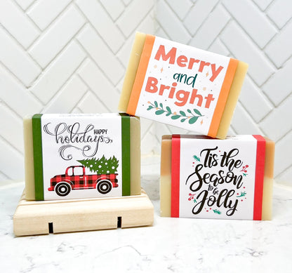 Three holiday-themed bar soaps with decorative labels displayed on a light surface, one resting on a wooden soap dish.