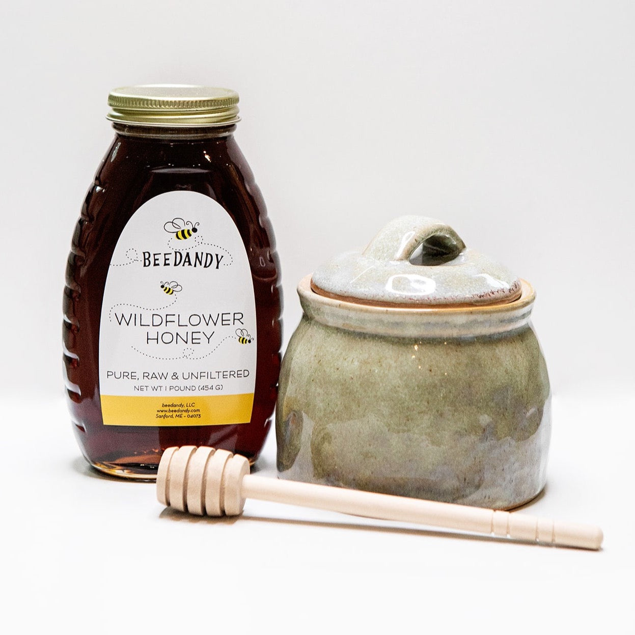 Jar of Beedandy wildflower honey displayed beside a ceramic honey pot and a wooden honey dipper on a white background.