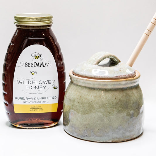 Jar of Beedandy wildflower honey displayed beside a ceramic honey pot with a wooden honey dipper sticking out, on a white background.