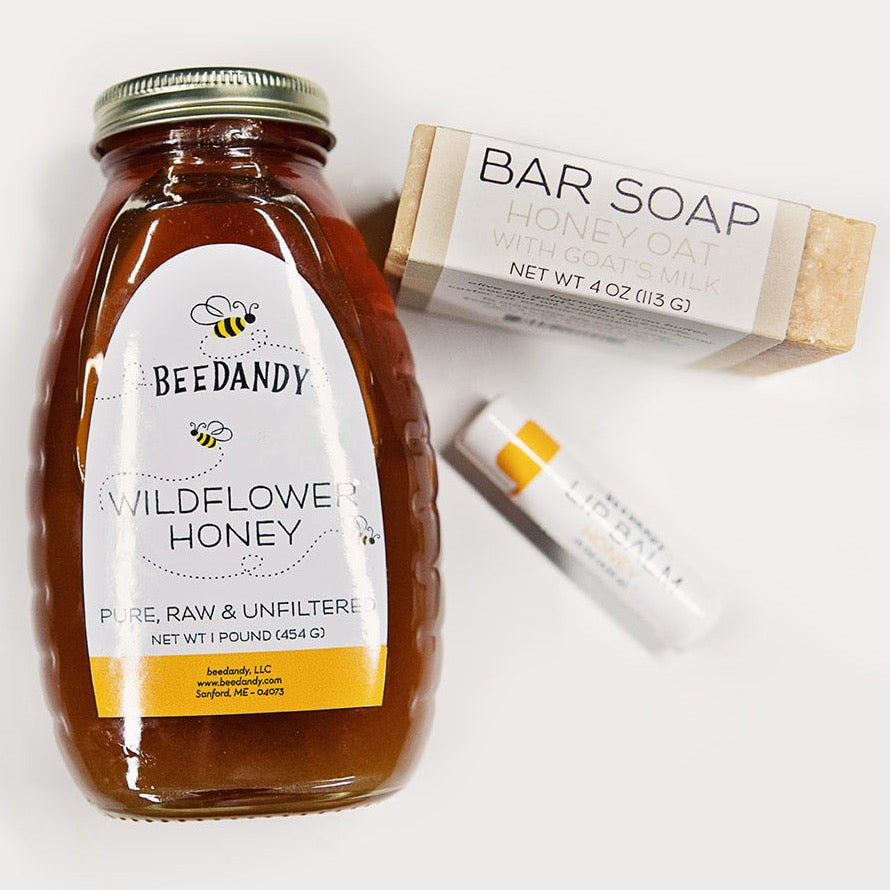 A gift set containing a jar of wildflower honey, a bar of honey oat soap, and a tube of honey lip balm, all branded with 'BEEDANDY'. The items are arranged in front of a white background.