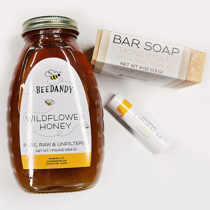 A gift set containing a jar of wildflower honey, a bar of honey oat soap, and a tube of honey lip balm, all branded with 'BEEDANDY'. The items are arranged in front of a white background.