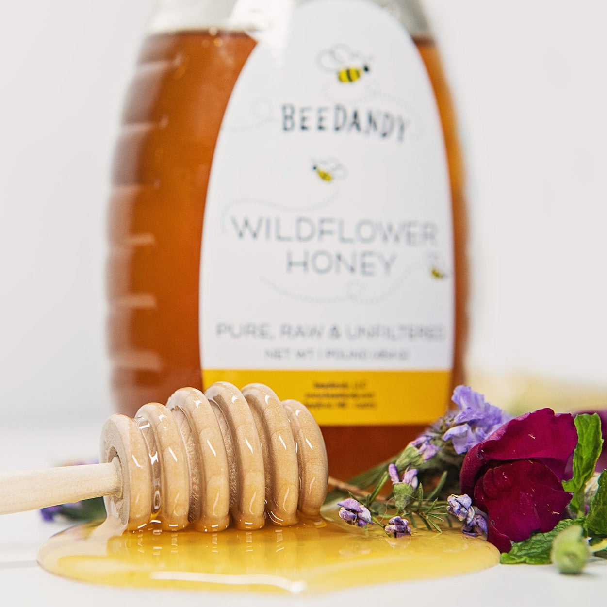 Wooden honey dipper resting in a pool of honey with a bottle of Beedandy wildflower honey in the background.