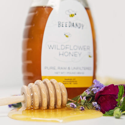 Wooden honey dipper resting in a pool of honey with a bottle of Beedandy wildflower honey in the background.