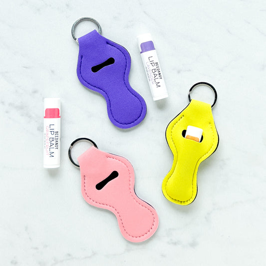 Neoprene lip balm holders in pink, yellow, and purple colors with a metal keyring and clip, displayed alongside lip balm tubes.