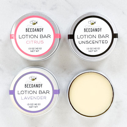 Four round metal tins of Beedandy lotion bars, including citrus, unscented, and lavender varieties, shown from above, with one tin open to display the lotion bar.