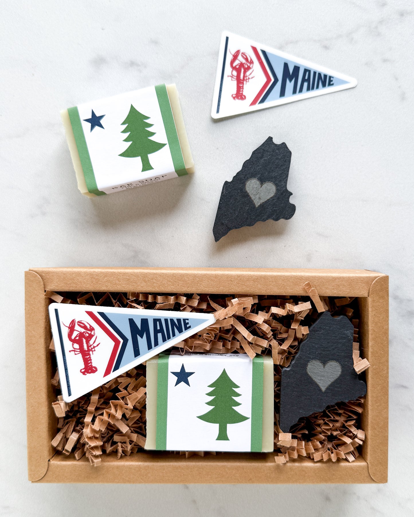 A gift box containing Maine-themed items, including a Maine pennant, a wrapped bar with a pine tree design, and a slate Maine-shaped ornament, shown from above with one box open and one closed. Another set of items beside box on white background.