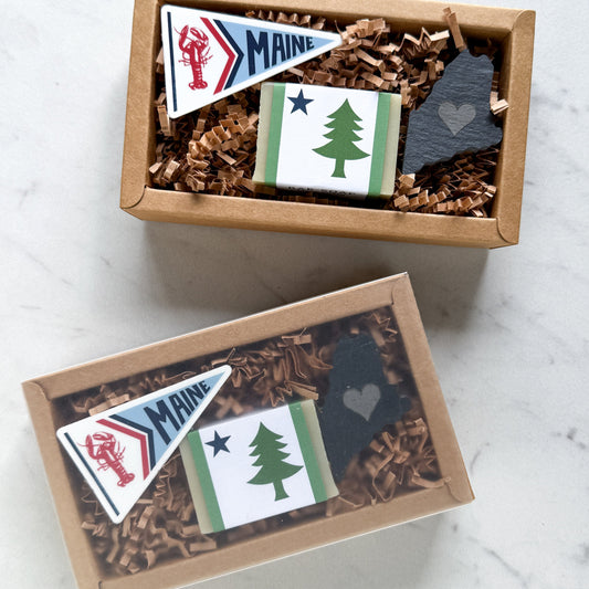 Two gift boxes containing Maine-themed items, including a Maine pennant sticker, a wrapped soap bar with a pine tree design, and a slate Maine-shaped magnet, shown from above with one box open and one closed.