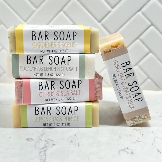 A collection of bar soaps with various labels indicating different scents and types, placed on a marble surface with white tile wall in the background.