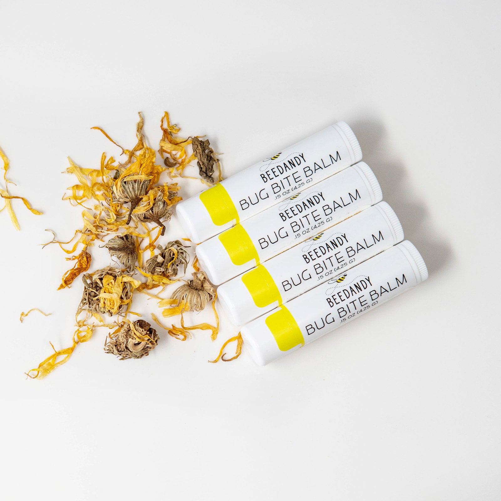 Four small white tubes with yellow labeling labeled Bug Bite Balm. All on a white background. 4 tubes are lined up with each other in a line with a small pile of calendula flowers. 