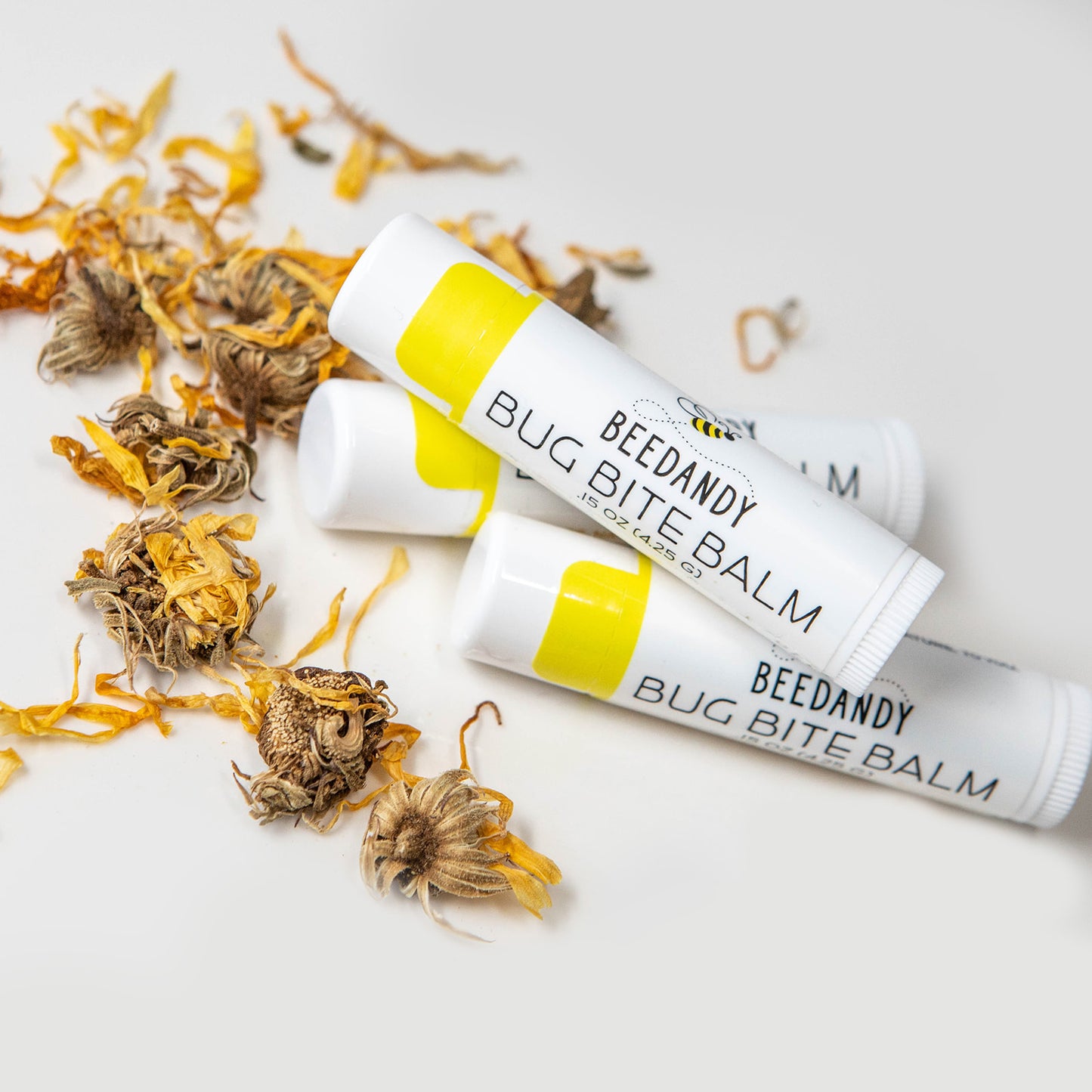 Three small white tubes with yellow labeling labeled Bug Bite Balm. 3 tubes are stacked up (one on top of other two) with a small pile of calendula flowers. All on a white background. 