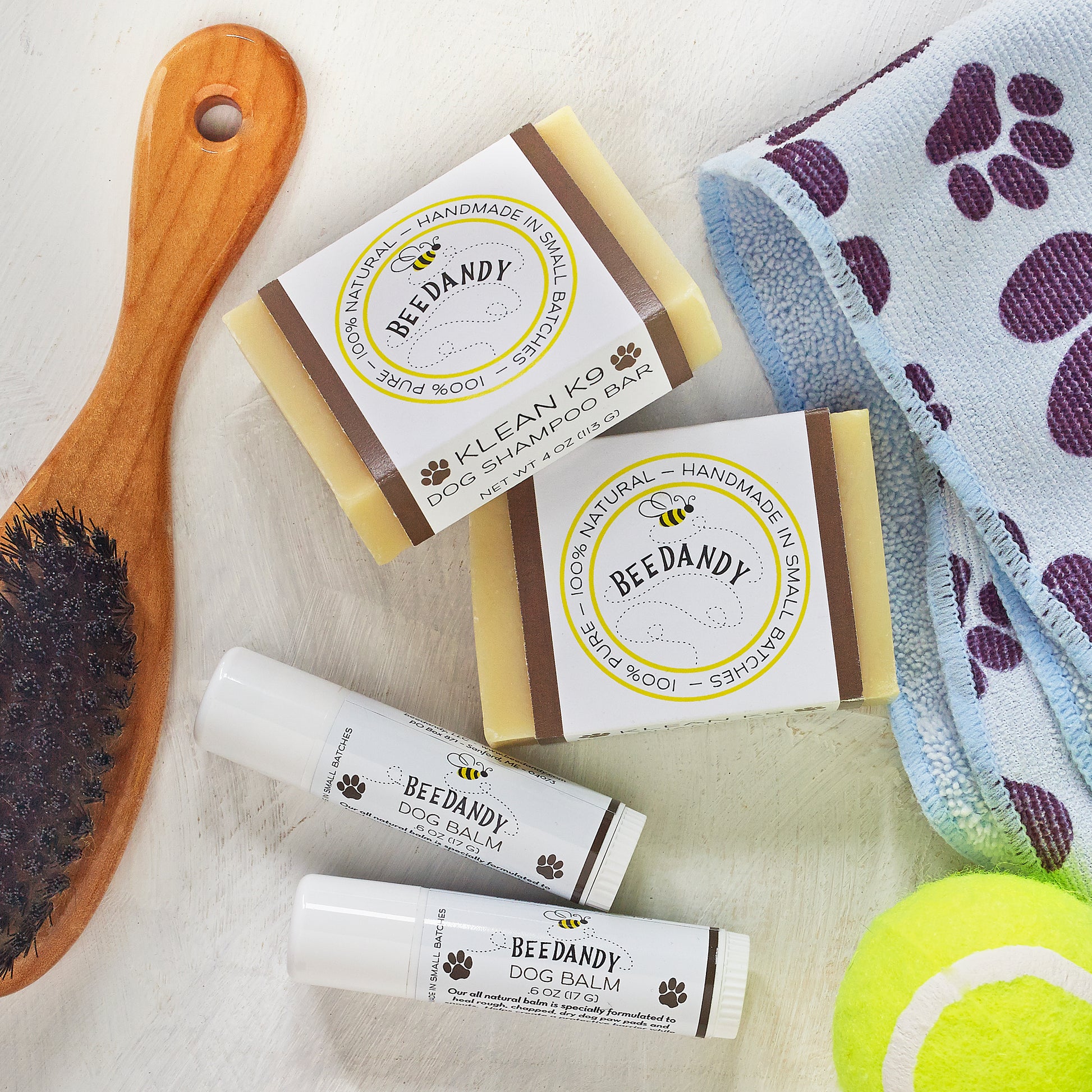 Beedandy dog shampoo bars and dog balm tubes arranged with a wooden brush, towel, and tennis ball on a light surface.